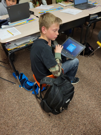 This boy's backpack is bigger than the chair he's sitting in!