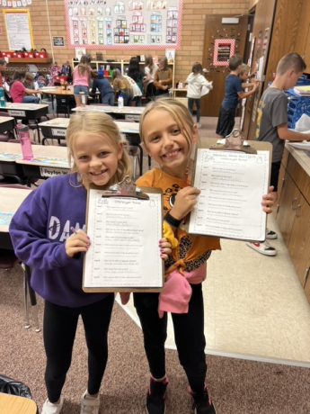 2 girls smile while showing their work on clipboards